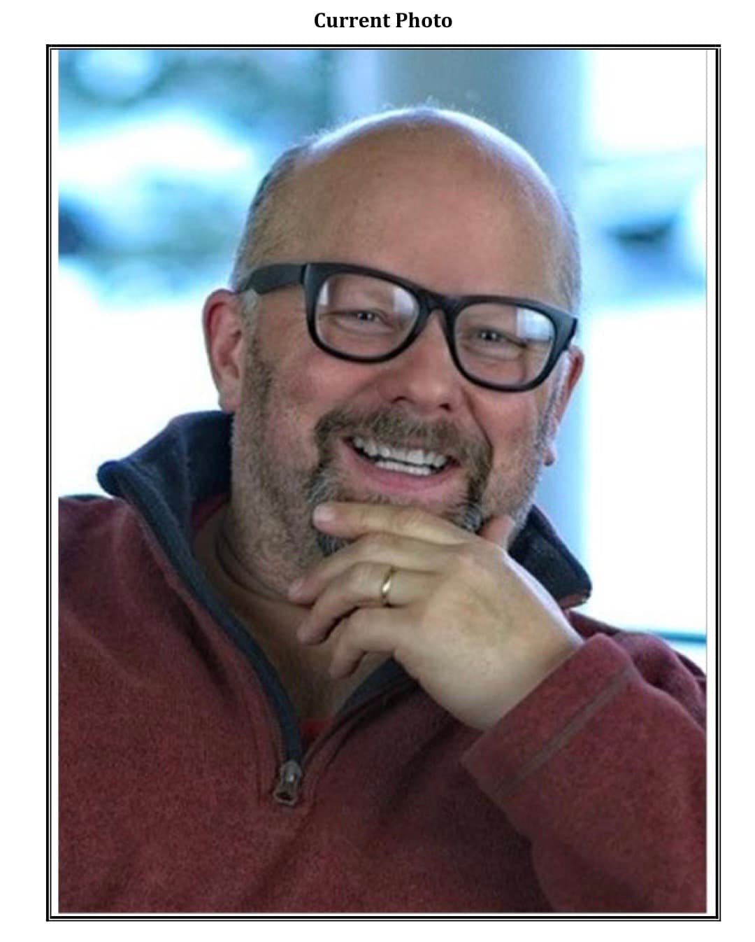 Smiling bald man with glasses and beard, wearing red fleece, resting hand on chin.